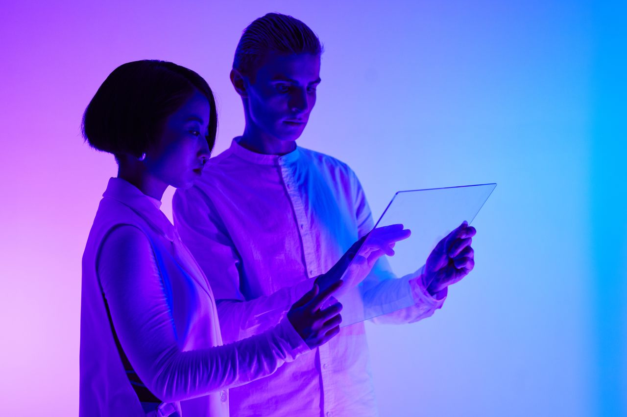 services-01 Two adults engaging with transparent screen, illuminated by vibrant purple and blue lights.